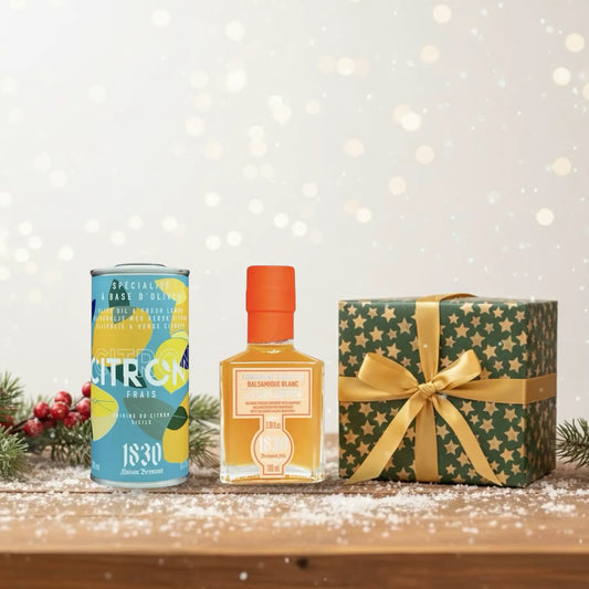 Geschenk mit goldener Schleife, Olivenöl mit Zitrone und Grapefruit-Balsam-Essig von Maison Brémond 1830, festlich dekoriert auf verschneitem Holztisch mit Tannenzweig und roten Beeren im Hintergrund.
