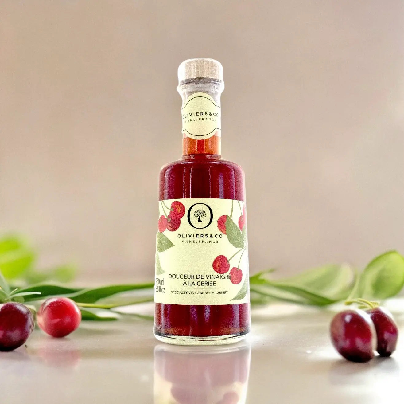 eine flasche kirschessig von oliviers & co steht auf einer reflektierenden oberfläche. die flasche hat eine etikette mit roten kirschen und grünen blättern. im hintergrund liegen grüne blätter und zwei frische kirschen