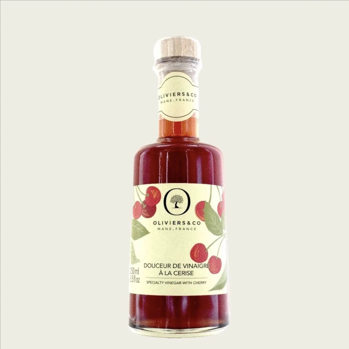 eine flasche kirschessig von oliviers & co steht auf einer weißen oberfläche. die flasche hat eine etikette mit roten kirschen und grünen blättern. im hintergrund sind unscharfe grüne blätter zu sehen