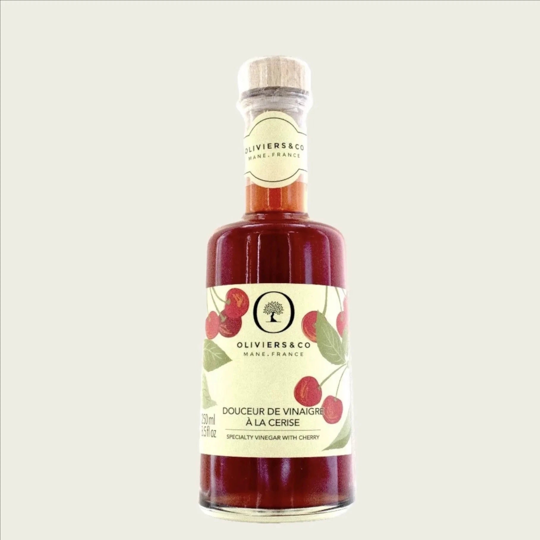eine flasche kirschessig von oliviers & co steht auf einer weißen oberfläche. die flasche hat eine etikette mit roten kirschen und grünen blättern. im hintergrund sind unscharfe grüne blätter zu sehen