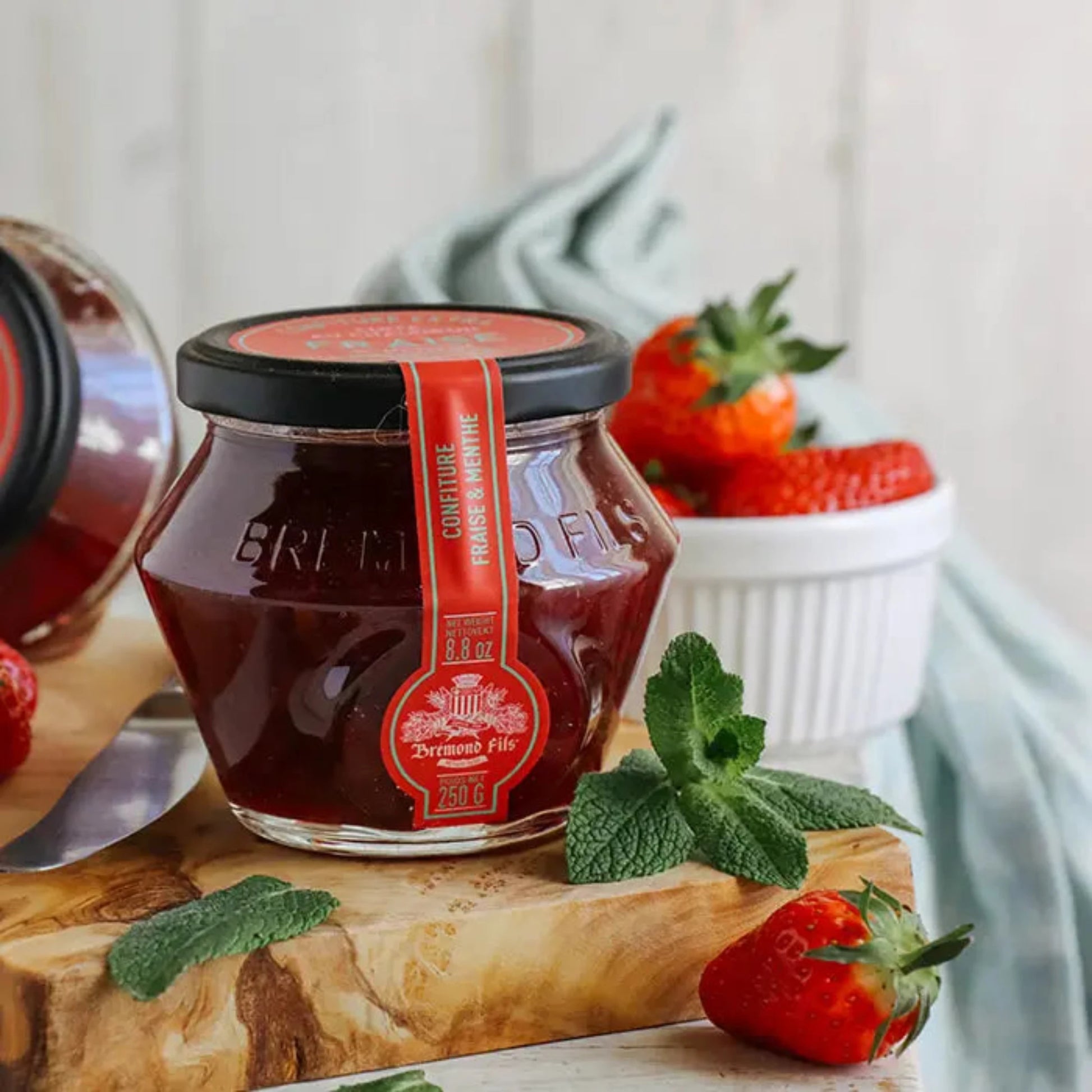 Erdbeer-Minze-Konfitüre im Glas mit rotem Etikett von Maison Brémond, dekoriert mit frischen Erdbeeren und Minzblättern auf einem Holzbrett.