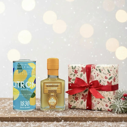 Geschenk mit roter Schleife, Olivenöl mit Zitrone und Mango-Balsam-Essig von Maison Brémond 1830, festlich arrangiert auf verschneitem Holztisch vor winterlichem Hintergrund.