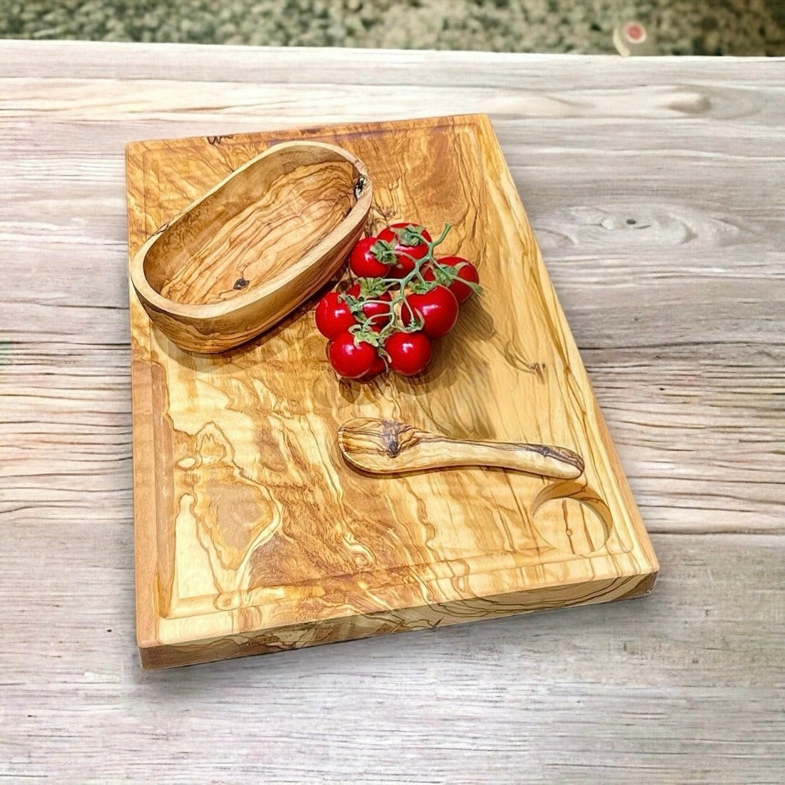 Ein rechteckiges Schneidebrett aus Olivenholz mit einer natürlichen Maserung, platziert auf einem Holztisch. Auf dem Brett befinden sich eine kleine Holzschale, ein Holzlöffel und eine Rispe mit roten Tomaten.