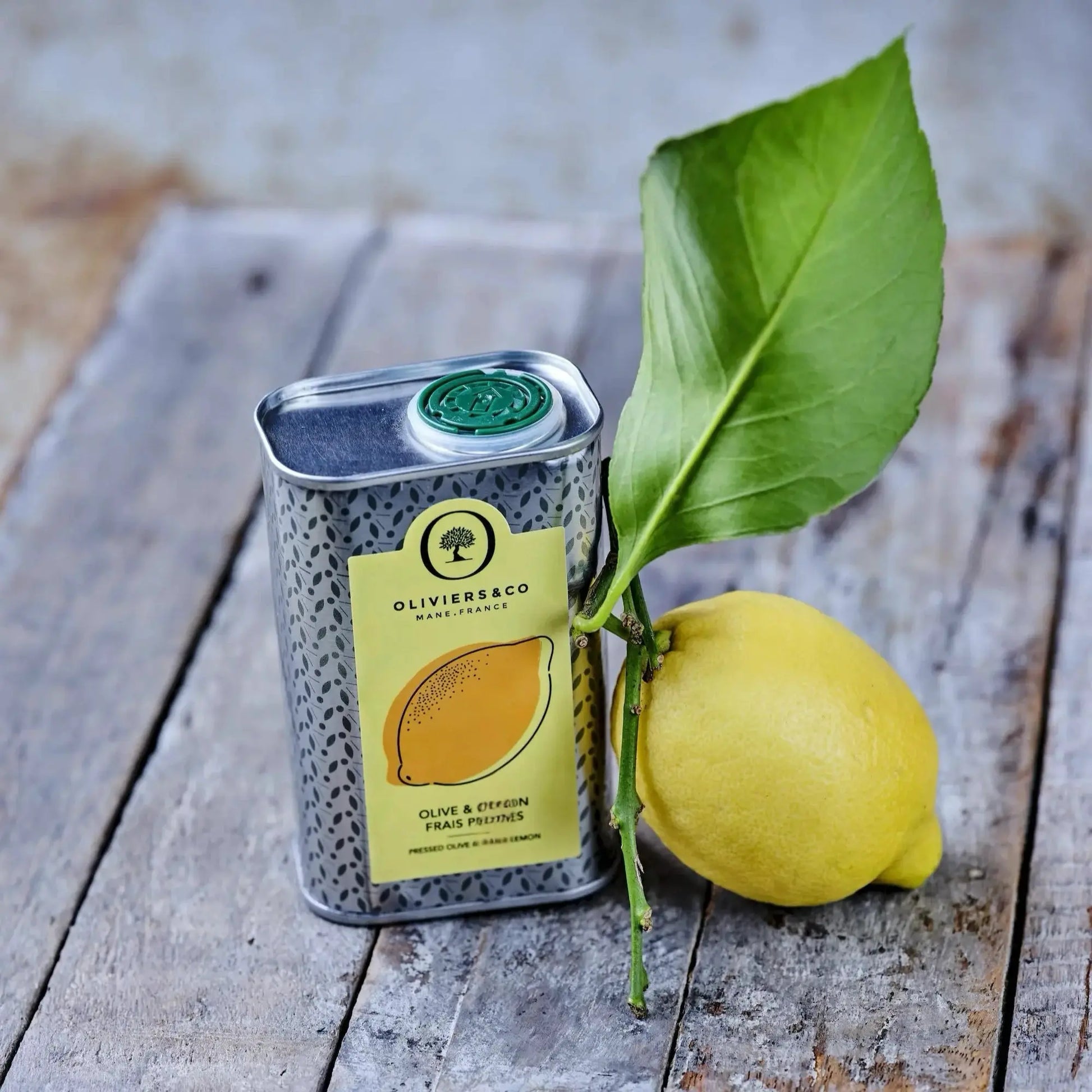 Eine Dose Bio-Olivenöl mit Zitronengeschmack steht auf einem rustikalen Holzbrett. Auf der Dose ist ein Etikett mit der Aufschrift „Olive & Citron Frais Pressés“ zu sehen. Neben der Dose liegt eine ganze Zitrone mit einem grünen Blatt, die an der Dose lehnt, und ein Stück abgeschnittene Zitrone liegt vorne links im Bild. Der Hintergrund ist unscharf und zeigt eine blaugrüne, verwitterte Oberfläche. 