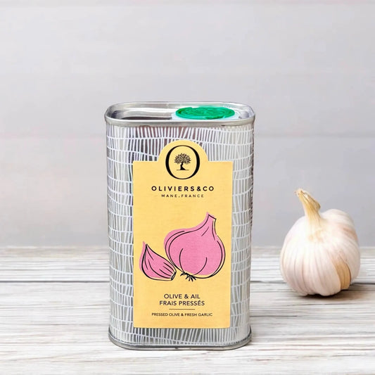 olivenoel mit knoblauch. ein rechteckiger metallbehälter mit einem gelben etikett, auf dem zwei rosa knoblauchknollen abgebildet sind. darauf steht "oliviers & co mane, france" und "olive & ail frais pressés". neben dem behälter liegt eine einzelne knoblauchknolle auf einer hellen, strukturierten oberfläche. der hintergrund ist schlicht und hellgrau.