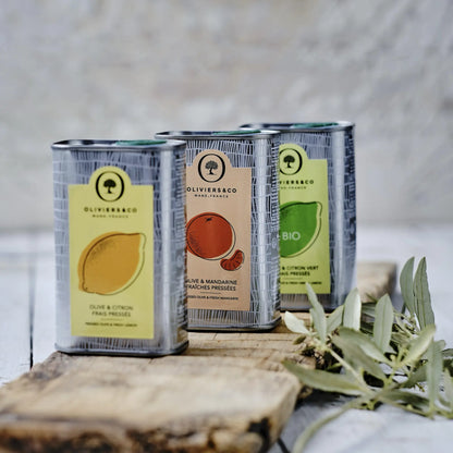 drei dosen olivenöl von oliviers&co, eine mit zitrone, eine mit mandarine und eine mit grüner zitrone, auf einem holzbrett, mit olivenzweigen daneben
