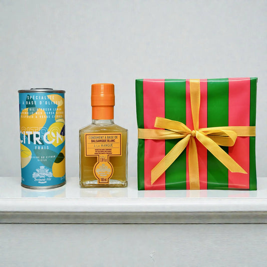 Geschenkset mit Zitronen-Olivenöl in Dose, Mango-Balsamico-Condiment in Glasflasche und dekorativer Geschenkverpackung mit Schleife auf weißem Regal
