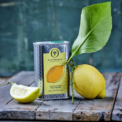 Eine Dose Bio-Olivenöl mit Zitronengeschmack steht auf einem rustikalen Holzbrett. Auf der Dose ist ein Etikett mit der Aufschrift „Olive & Citron Frais Pressés“ zu sehen. Neben der Dose liegt eine ganze Zitrone mit einem grünen Blatt, die an der Dose lehnt, und ein Stück abgeschnittene Zitrone liegt vorne links im Bild. Der Hintergrund ist unscharf und zeigt eine blaugrüne, verwitterte Oberfläche. 