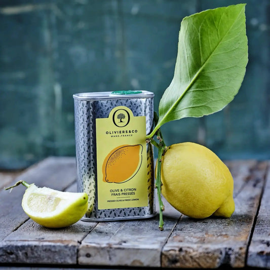 Eine Dose Bio-Olivenöl mit Zitronengeschmack steht auf einem rustikalen Holzbrett. Auf der Dose ist ein Etikett mit der Aufschrift „Olive & Citron Frais Pressés“ zu sehen. Neben der Dose liegt eine ganze Zitrone mit einem grünen Blatt, die an der Dose lehnt, und ein Stück abgeschnittene Zitrone liegt vorne links im Bild. Der Hintergrund ist unscharf und zeigt eine blaugrüne, verwitterte Oberfläche. 