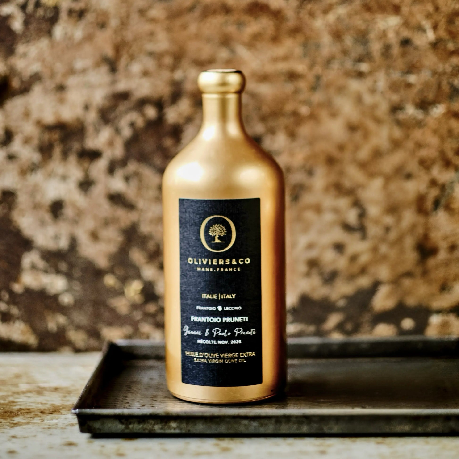 Goldene Flasche Olivenöl aus Italien der Marke Oliviers & Co, präsentiert auf einem dunklen Tablett vor einer rustikalen Steinwand. Das schwarze Etikett zeigt die Bezeichnung "Frantoio Pruneti" und "Extra Virgin Olive Oil", mit Details zur Herkunft aus Italien und dem Erntedatum im November 2023. Die hochwertige Verpackung und die edle Präsentation unterstreichen die Exklusivität des Olivenöls.