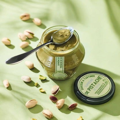 Pistaziencreme-Glas mit einem Löffel Pistaziencreme, dekorativ arrangiert auf grünem Hintergrund mit Pistazien.
