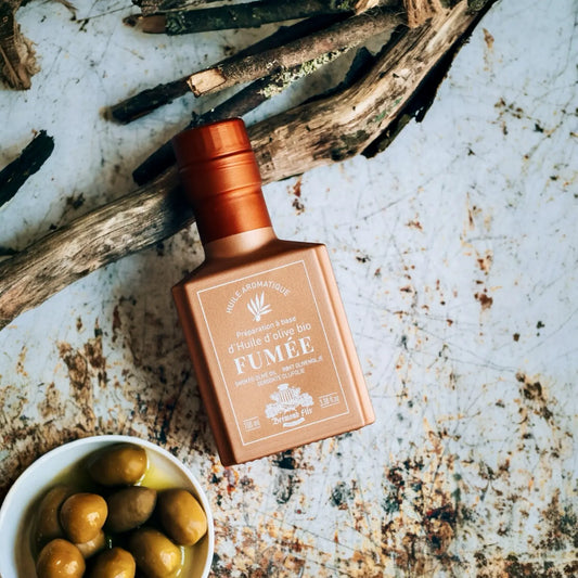 Bronzefarbene Flasche mit rauchigem Bio-Olivenöl der Marke 'Deromé Fils', umgeben von rustikalen Holzstücken und einer Schale mit grünen Oliven, auf einer verwitterten Metalloberfläche.