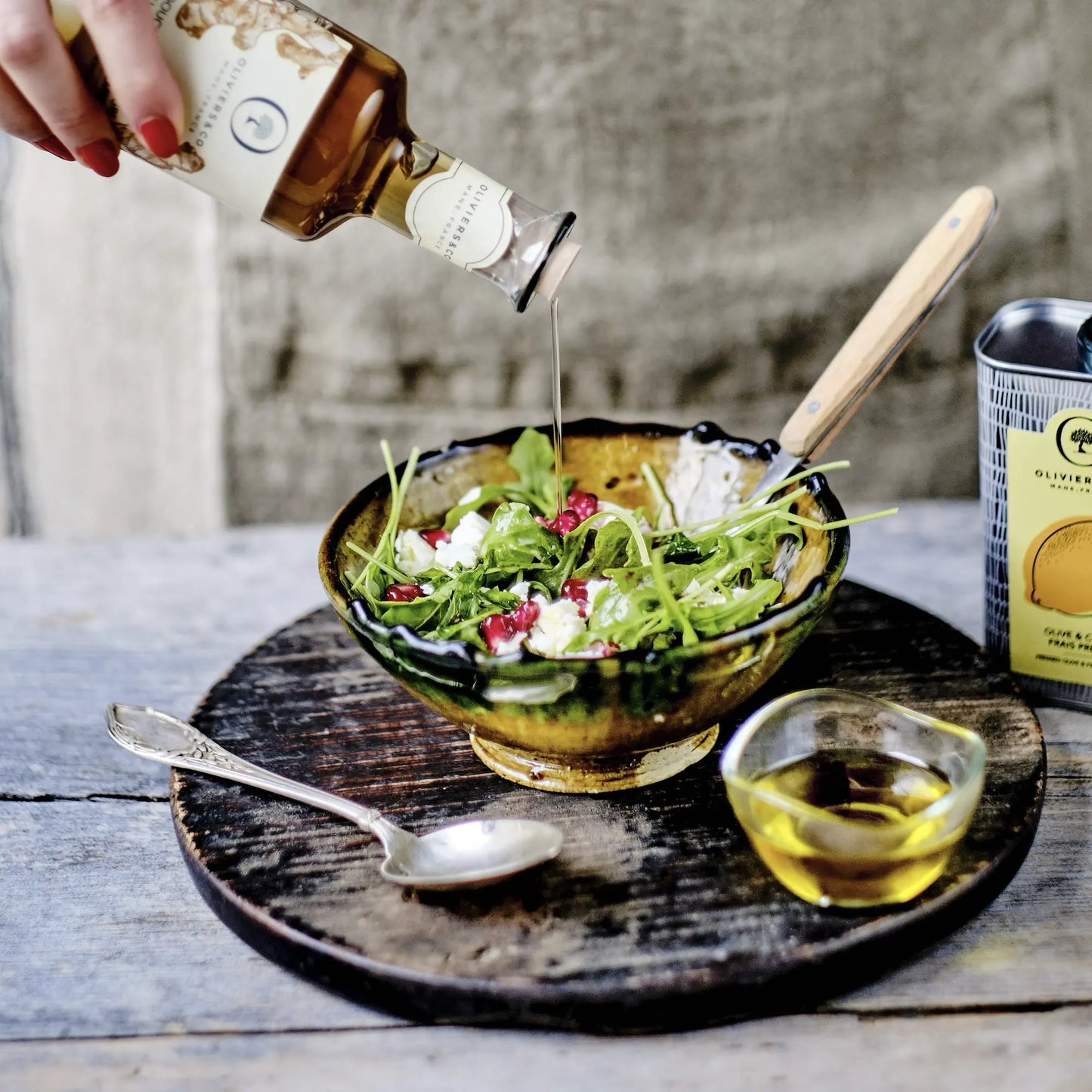 eine hand mit roten nägeln gießt olivenöl von oliviers&co auf einen salat mit granatapfelkernen und feta in einer braunen schüssel, daneben stehen eine dose olivenöl mit zitrone und eine kleine schale mit öl