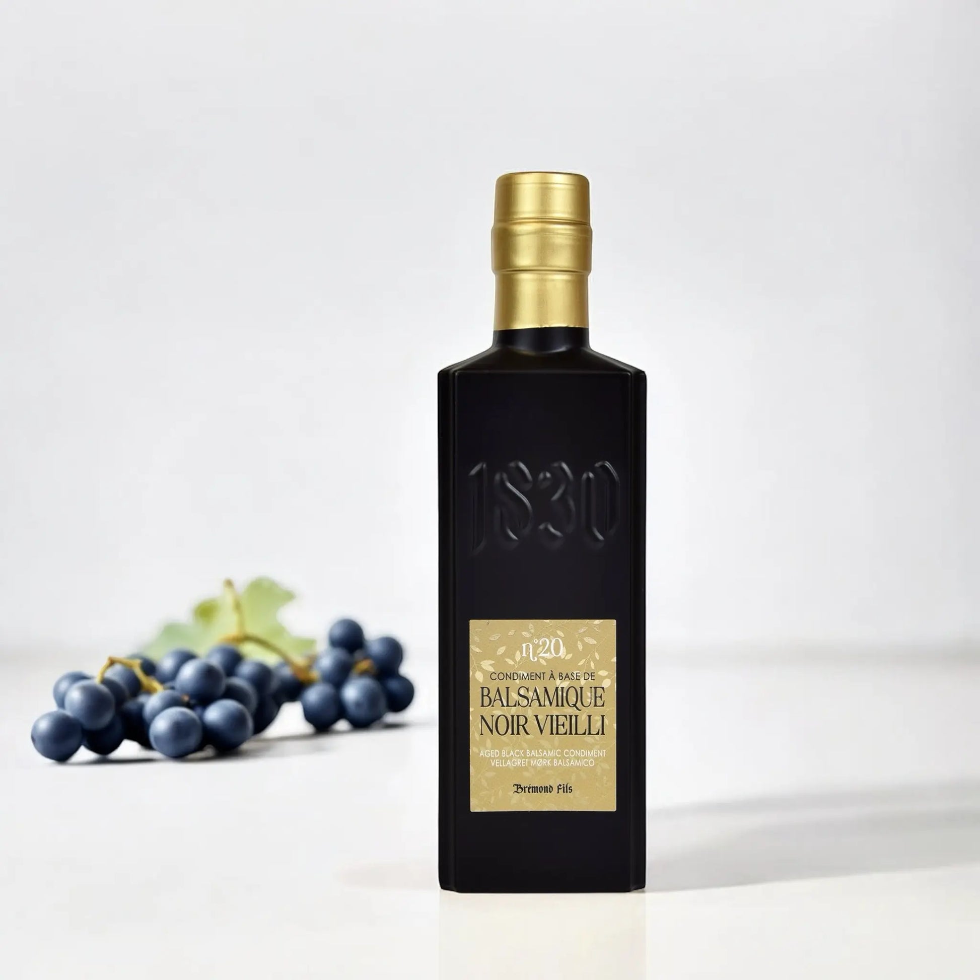 Hochwertiger schwarzer Balsamico-Essig – 20 Jahre gereift, präsentiert mit frischen Weintrauben auf weißem Hintergrund – ein Zeichen für höchste Qualität.
