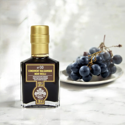 Edler schwarzer Balsamico-Essig – 30 Jahre gereift in einer hochwertigen Glasflasche, mit dunklen Trauben im Hintergrund.