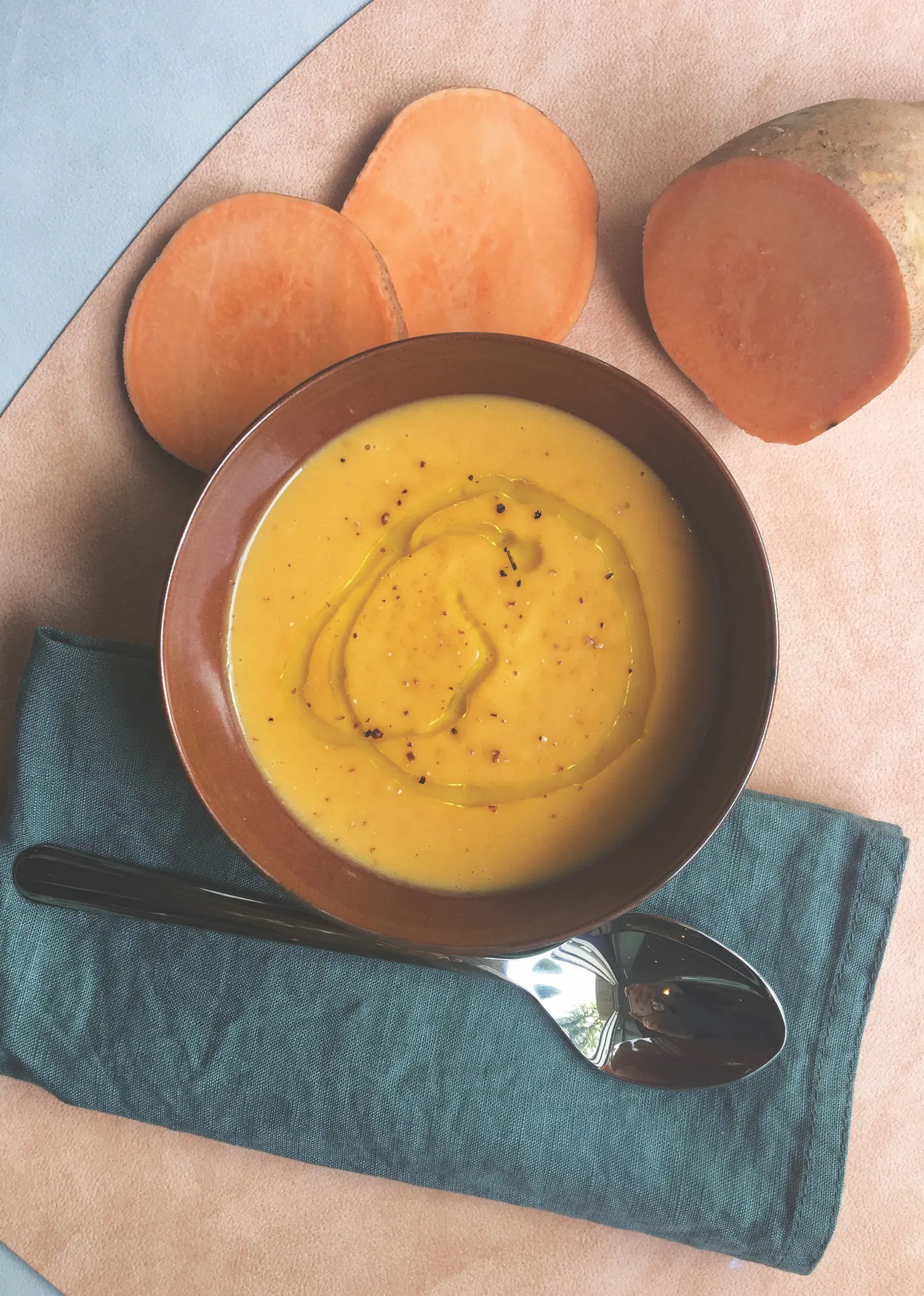 Cremige Süßkartoffelsuppe, garniert mit einem Schuss Trüffel Olivenöl, serviert in einer braunen Schale. Frische Süßkartoffelscheiben und ein eleganter Löffel auf einer Stoffserviette ergänzen das Arrangement. Perfekt für herzhafte Rezepte mit hochwertigen Zutaten.