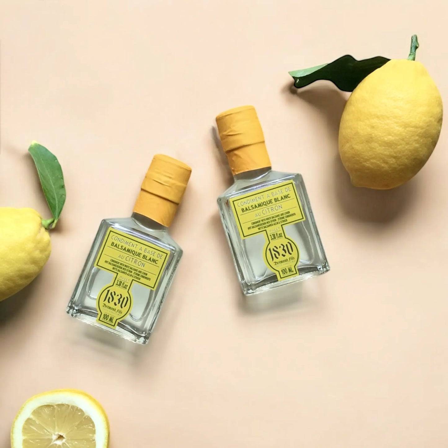 Zwei Flaschen weißer Balsamico Essig mit Zitrone, 100 ml, mit gelbem Verschluss, dekoriert mit frischen Zitronen und einer halben Zitrone auf hellem Hintergrund. Perfekt für kreative und fruchtige Gerichte.