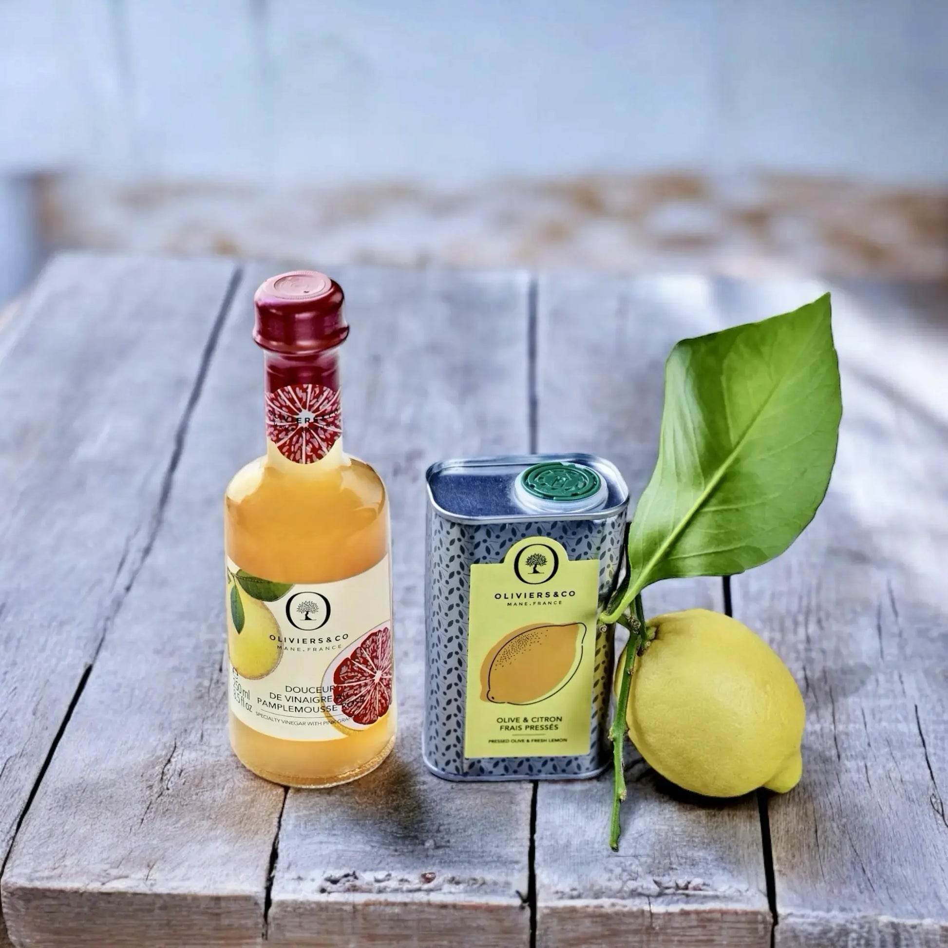 Eine Dose Bio-Olivenöl mit Zitrone und eine Flasche Grapefruitessig stehen auf einem rustikalen Holztisch, begleitet von einer frischen Zitrone mit Blatt, perfekt arrangiert für eine ansprechende Präsentation
